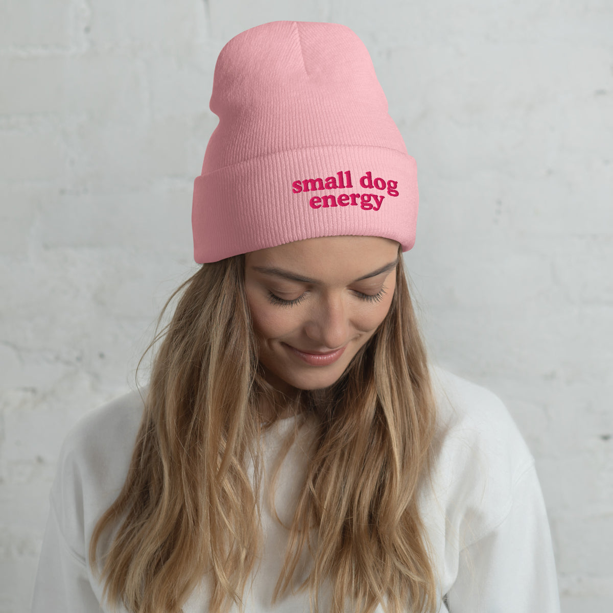 A woman wearing a light pink ribbed beanie with small dog energy embroidered on the front, paired with a white sweatshirt against a light gray brick wall.