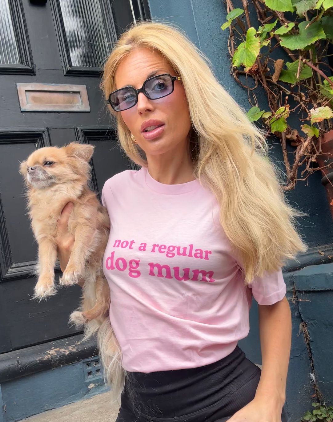 A customer in sunglasses wearing the pink Not a Regular Dog Mum tee while holding a small tan dog outside.