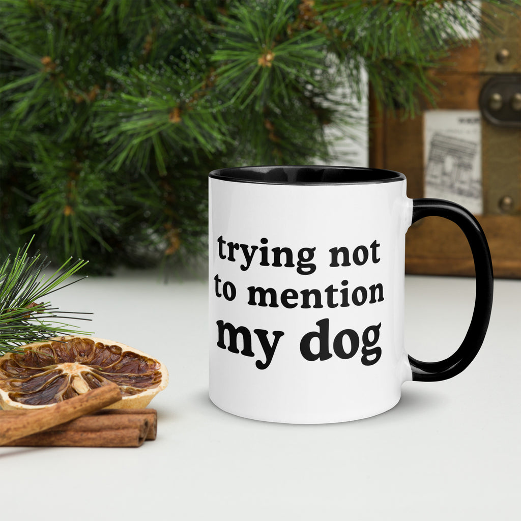 A white ceramic mug with a black interior and handle, printed with the phrase 'trying not to mention my dog' in black lowercase letters. The mug is styled on a tabletop with a dried orange slice, cinnamon sticks, and green pine branches in the background.