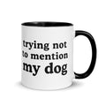 A white ceramic coffee mug with a black handle and black interior, featuring the humorous text 'trying not to mention my dog' printed in black lowercase letters on the front.