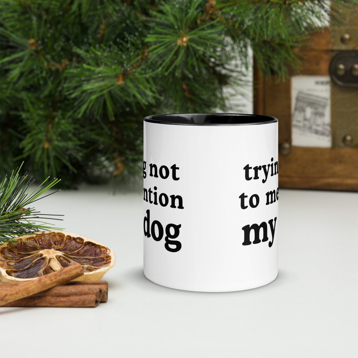 A white ceramic mug with a black interior and black printed text, styled on a tabletop with pine branches, a dried orange slice, cinnamon sticks, and a wooden box in the background.